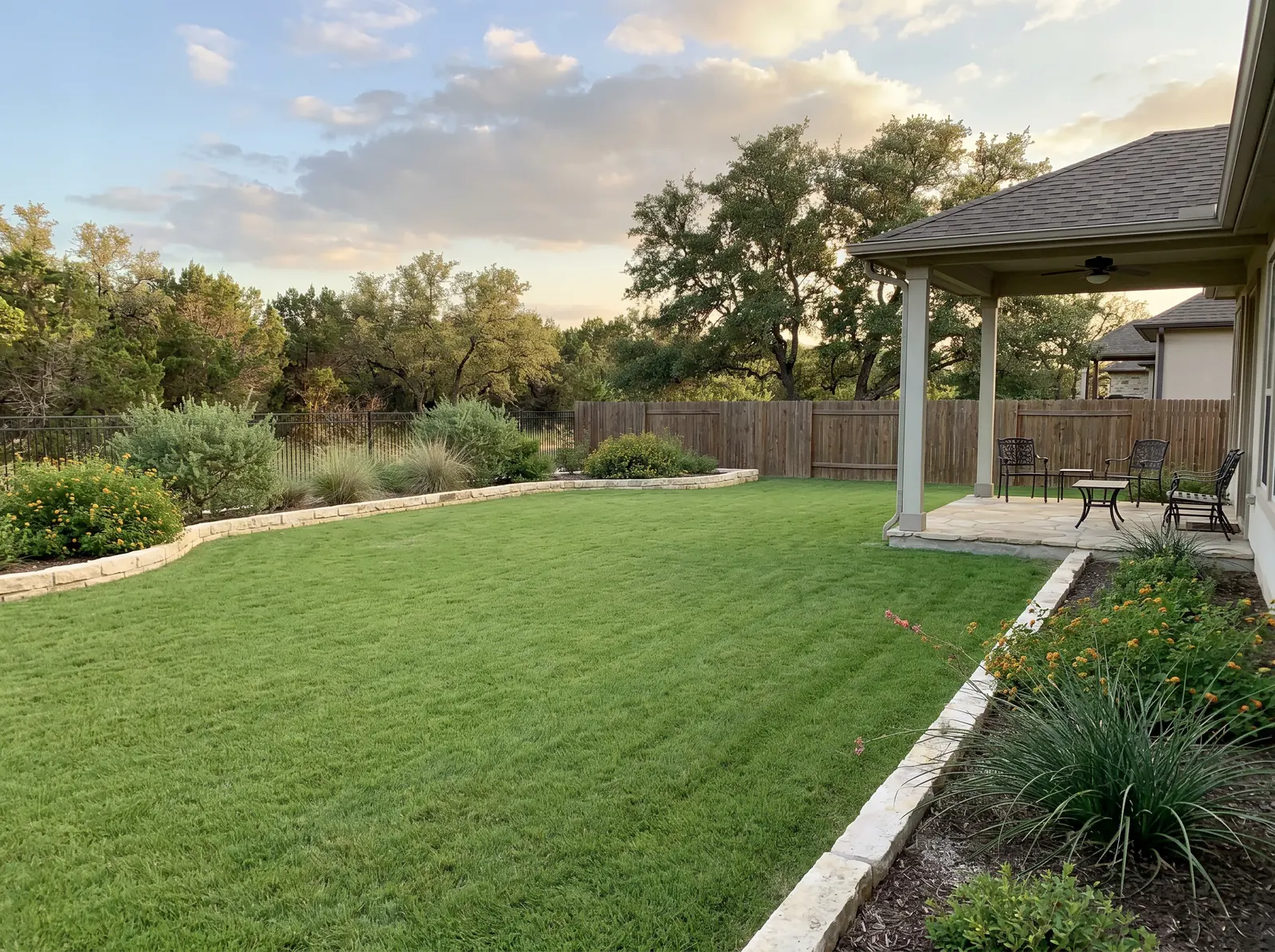 Backyard with green lawn, patio, curved stone border, and native shrubs at sunset.