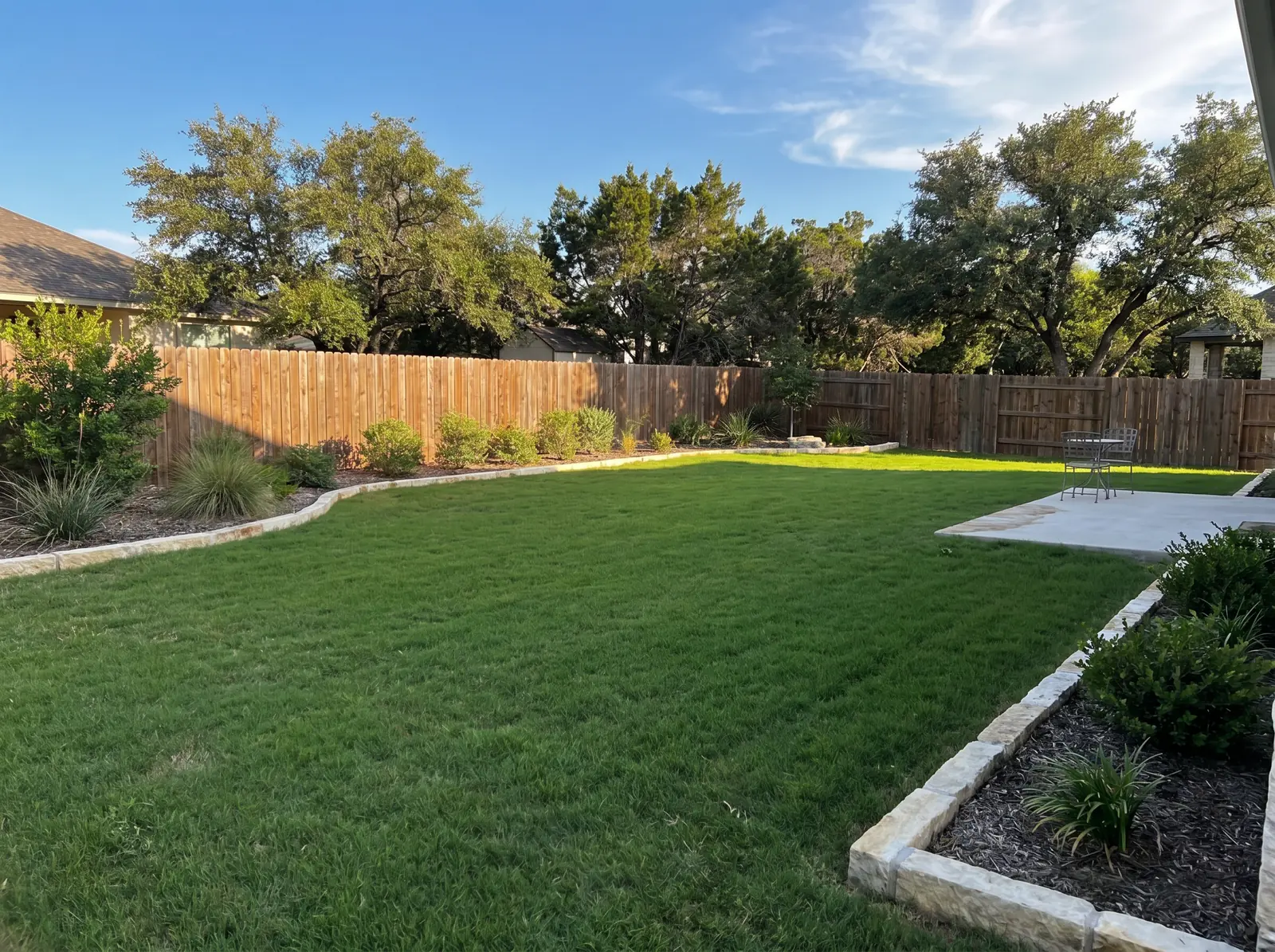 Backyard lawn bordered by stone edging, planting beds, wood fence, and patio.